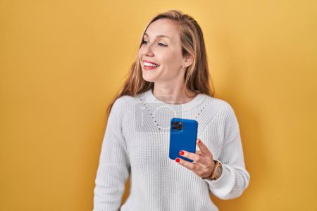 Photo for Young blonde woman using smartphone typing message looking away to side with smile on face, natural expression. laughing confident. - Royalty Free Image