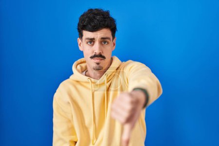 Photo for Hispanic man standing over blue background looking unhappy and angry showing rejection and negative with thumbs down gesture. bad expression. - Royalty Free Image