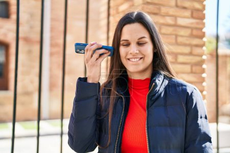 Téléchargez les photos : Jeune belle femme hispanique souriant confiant écoute message audio par le smartphonee à la rue - en image libre de droit