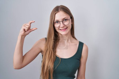Téléchargez les photos : Jeune femme caucasienne debout sur fond blanc geste souriant et confiant avec la main faisant signe de petite taille avec les doigts regardant et la caméra. concept de mesure. - en image libre de droit