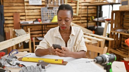 Foto de Carpintero femenino afroamericano serio que envía mensajes de texto hábilmente en el teléfono inteligente mientras se sienta con gracia en la mesa del taller de carpintería - Imagen libre de derechos