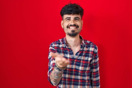 Foto de Joven hombre hispano con barba de pie sobre fondo rojo sonriente alegre ofreciendo mano de palma dando asistencia y aceptación. - Imagen libre de derechos