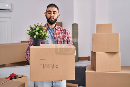 Photo for Middle east man with beard moving to a new home holding cardboard box making fish face with mouth and squinting eyes, crazy and comical. - Royalty Free Image