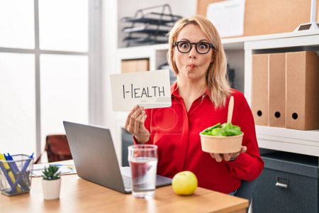 Photo for Blonde woman working at the office eating healthy food making fish face with mouth and squinting eyes, crazy and comical. - Royalty Free Image
