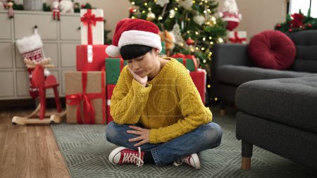 Téléchargez les photos : Jeune femme chinoise assise près d'un arbre de Noël avec une expression ennuyeuse à la maison - en image libre de droit