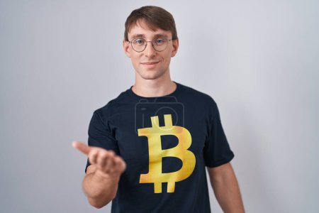 Téléchargez les photos : Blanc blond homme portant Bitcoin t-shirt souriant gai offrant main de paume donnant assistance et acceptation. - en image libre de droit