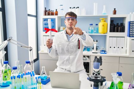 Photo for Young hispanic man working at scientist laboratory holding diploma pointing with finger to the camera and to you, confident gesture looking serious - Royalty Free Image