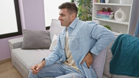 Portrait of worried, unhappy, young hispanic man sitting on sofa at home, seriously suffering from intense backache pain, indoor living room background
