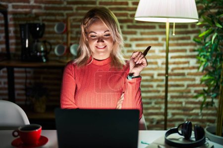 Photo for Blonde woman using laptop at night at home with a big smile on face, pointing with hand and finger to the side looking at the camera. - Royalty Free Image