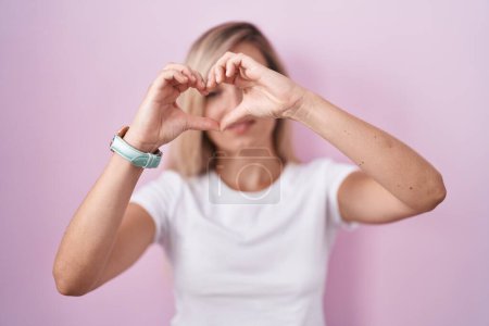 Téléchargez les photos : Jeune femme blonde debout sur fond rose faisant forme de coeur avec la main et les doigts souriant en regardant à travers signe - en image libre de droit
