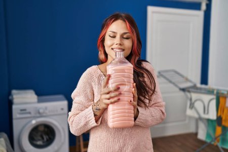 Foto de Joven mujer caucásica sonriendo confiado oliendo botella de detergente en la sala de lavandería - Imagen libre de derechos