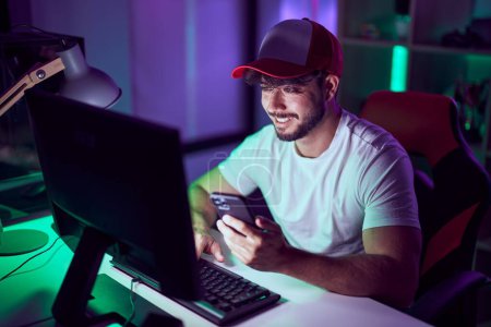 Téléchargez les photos : Jeune homme hispanique streamer en utilisant l'ordinateur et smarpthone à la salle de jeu - en image libre de droit