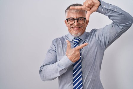 Foto de Hombre de negocios hispano de pelo gris con gafas sonrientes haciendo montura con las manos y los dedos con la cara feliz. concepto de creatividad y fotografía. - Imagen libre de derechos