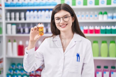 Foto de Joven mujer caucásica farmacéutico sonriendo confiado sosteniendo pastillas botella en la farmacia - Imagen libre de derechos