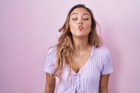 Foto de Mujer hispana joven de pie sobre fondo rosa haciendo cara de pez con labios, gesto loco y cómico. expresión divertida. - Imagen libre de derechos