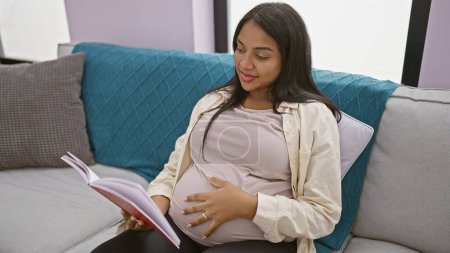 Téléchargez les photos : Brillante jeune femme enceinte jouissant d'un repos confortable, absorbée dans le livre de lecture tout en touchant le ventre et rayonnant de positivité à la maison - en image libre de droit