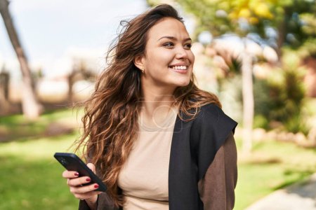 Foto de Joven hermosa mujer hispana sonriendo confiada usando teléfono inteligente en el parque - Imagen libre de derechos