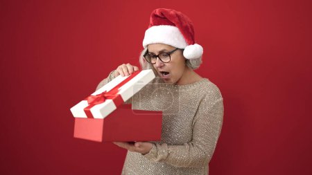 Téléchargez les photos : Femme d'âge moyen avec des cheveux gris portant un chapeau de Noël déballer cadeau avec visage surpris sur fond rouge isolé - en image libre de droit