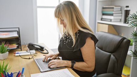 Photo for Serious yet relaxed, beautiful middle-aged blonde business woman successfully bossing around. focused, working on laptop at office desk against room's indoor background. - Royalty Free Image