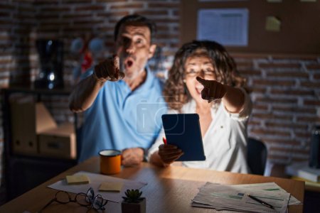 Photo for Middle age hispanic couple using touchpad sitting on the table at night pointing with finger surprised ahead, open mouth amazed expression, something on the front - Royalty Free Image