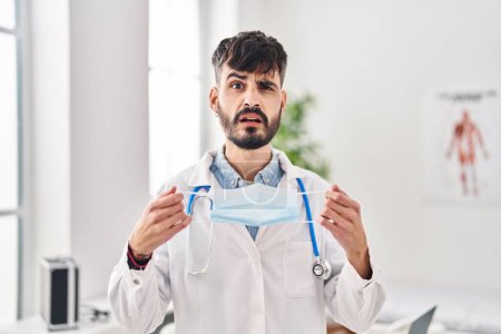 Photo for Young hispanic doctor man with beard holding safety mask clueless and confused expression. doubt concept. - Royalty Free Image