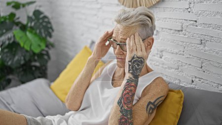 Photo for Stressed grey-haired senior woman suffering from a headache, sitting on the bed in her bedroom expressing worry and exhaustion - Royalty Free Image