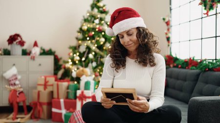 Foto de Mujer hispana de mediana edad mirando la foto sentada en el sofá por el árbol de Navidad en casa - Imagen libre de derechos