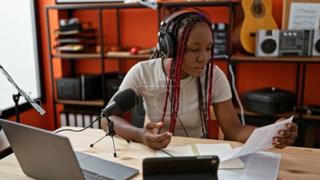 Foto de Reportera afroamericana confiada, transmitiendo noticias en vivo por la radio, leyendo periódicos en el estudio, auriculares encendidos - Imagen libre de derechos
