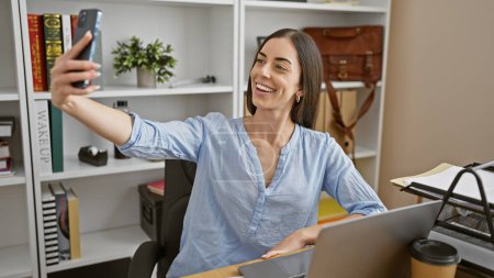Foto de Mujer hispana joven y radiante captura selfie con teléfono inteligente durante el trabajo de oficina, una instantánea de su exitoso estilo de vida empresarial en el elegante lugar de trabajo - Imagen libre de derechos