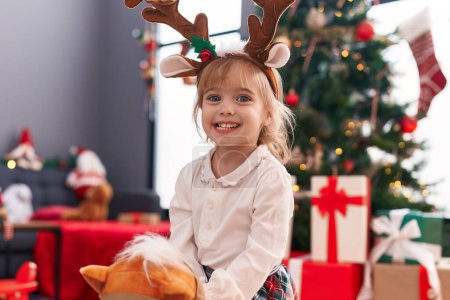 Photo for Adorable blonde girl playing with horse toy standing by christmas tree at home - Royalty Free Image
