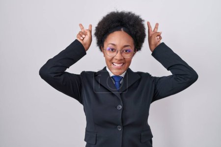 Foto de Hermosa mujer africana con el pelo rizado usando chaqueta de negocios y gafas posando divertido y loco con los dedos en la cabeza como orejas de conejo, sonriente alegre - Imagen libre de derechos