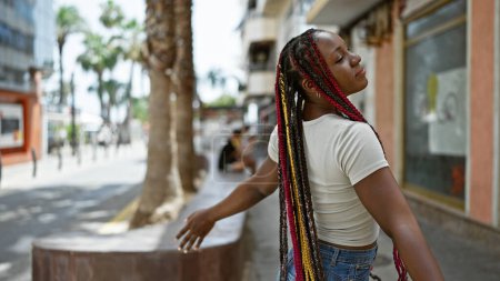 Téléchargez les photos : Belle femme afro-américaine avec des tresses, debout sur la rue ensoleillée, méditant les yeux fermés, respirant l'air de l'été, les bras grands ouverts, jouissant de sa liberté, immergée dans un état détendu. - en image libre de droit