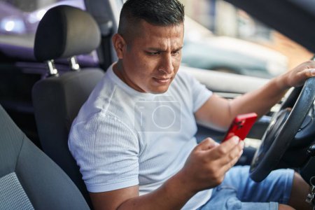 Téléchargez les photos : Jeune homme latin utilisant smartphone assis sur la voiture à la rue - en image libre de droit