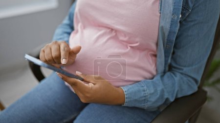 Photo for Young, pregnant businesswoman, embracing motherhood, relaxed, sitting at her office desk, busily texting on her smartphone, fully aware of her impending success. - Royalty Free Image