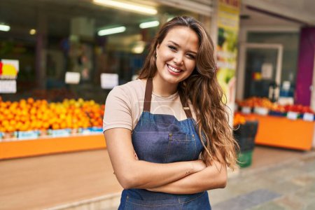 Téléchargez les photos : Jeune belle femme hispanique serveuse souriante confiante debout avec les bras croisés geste au magasin de fruits - en image libre de droit
