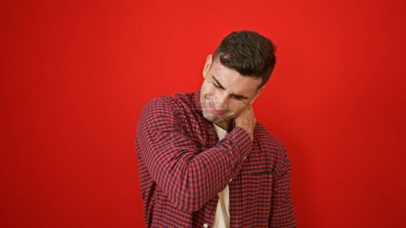 Serious young hispanic male suffering unbearable cervical back pain, standing alone over an isolated red background
