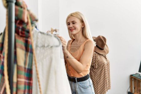 Foto de Joven mujer rubia cliente elegir ropa sonriendo en la tienda de ropa - Imagen libre de derechos