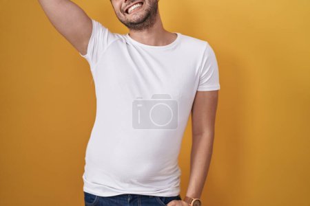 Photo for Hispanic man wearing white t shirt over yellow background stressed and frustrated with hand on head, surprised and angry face - Royalty Free Image
