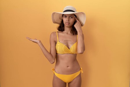 Foto de Mujer hispana joven con bikini y sombrero de verano confundida y molesta con la palma abierta que muestra espacio para copiar y señalar con el dedo a la frente. Piénsalo. - Imagen libre de derechos
