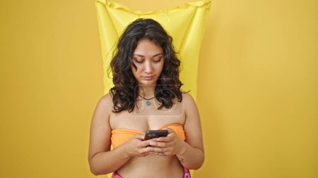 Téléchargez les photos : Young beautiful hispanic woman tourist wearing bikini using smartphone over mattress float over isolated yellow background - en image libre de droit