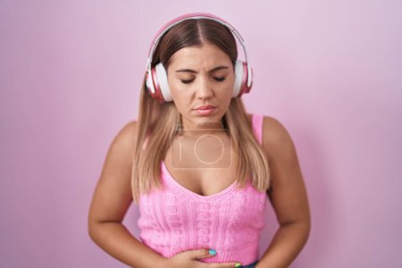 Photo for Young blonde woman listening to music using headphones with hand on stomach because indigestion, painful illness feeling unwell. ache concept. - Royalty Free Image