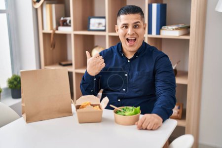 Photo for Hispanic young man eating take away food pointing thumb up to the side smiling happy with open mouth - Royalty Free Image