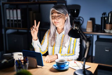 Téléchargez les photos : Femme d'âge moyen avec les cheveux gris travaillant au bureau la nuit montrant et pointant vers le haut avec les doigts numéro deux tout en souriant confiant et heureux. - en image libre de droit
