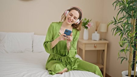 Foto de Young blonde woman listening to music sitting on bed at bedroom - Imagen libre de derechos