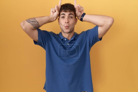 Photo for Young hispanic man standing over yellow background doing funny gesture with finger over head as bull horns - Royalty Free Image