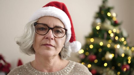 Téléchargez les photos : Femme d'âge moyen aux cheveux gris debout avec une expression sérieuse portant un chapeau de Noël à la maison - en image libre de droit