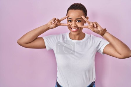 Foto de Hermosa mujer afroamericana de pie sobre fondo rosa haciendo símbolo de paz con los dedos sobre la cara, sonriendo alegre mostrando la victoria - Imagen libre de derechos