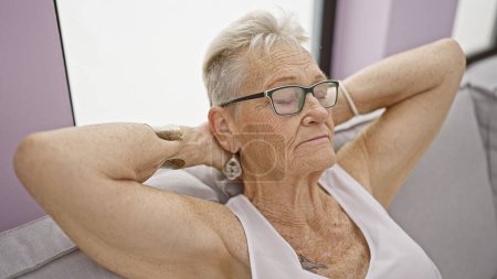 Téléchargez les photos : Femme âgée aux cheveux gris savoure un confort détendu, les mains sur la tête, assis sur le canapé à la maison, absorbant la lumière du soleil confortable dans la paix de son salon - en image libre de droit