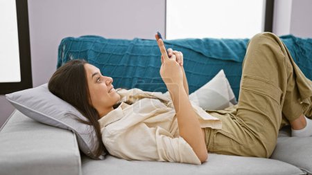 Foto de Joven mujer hispana alegre, tumbada en el sofá en casa, expresando sorpresa mientras usa su teléfono inteligente. - Imagen libre de derechos