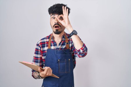 Téléchargez les photos : Jeune homme hispanique avec barbe portant tablier de serveur tenant presse-papiers faisant geste ok choqué par le visage surpris, oeil regardant à travers les doigts. expression incrédule. - en image libre de droit